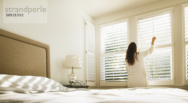 Frau im Bademantel öffnet Fensterläden im Schlafzimmer