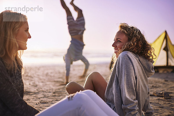 Lächelnde Frauen sprechen am Strand mit einem Mann  der im Hintergrund Handstand macht