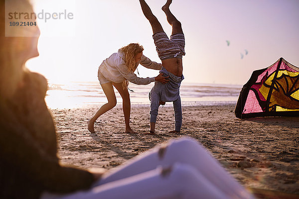 Frau hält Mann beim Handstand am Strand bei Sonnenuntergang