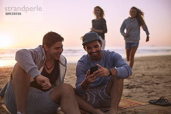 Freunde  die mit dem Handy am Strand bei Sonnenuntergang SMS schreiben.