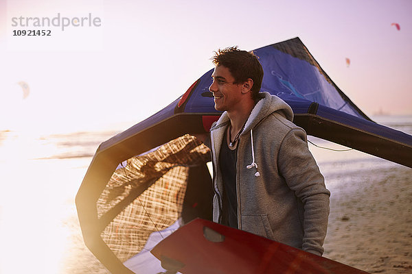 Lächelnder Mann mit Kiteboardausrüstung am Strand bei Sonnenuntergang