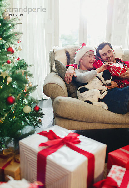 Paar Kuscheln auf Sofa mit Hund am Weihnachtsbaum und Geschenke