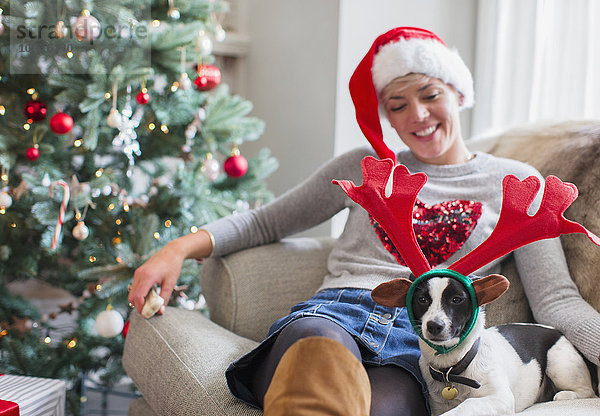 Frau auf Sofa sitzend mit Hund in Rentiergeweih am Weihnachtsbaum