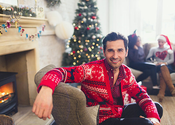 Portrait lächelnder Mann im Weihnachtspullover sitzend im Wohnzimmer