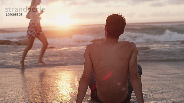 Junger Mann beobachtet Frau  die bei Sonnenuntergang in der Meeresbrandung läuft