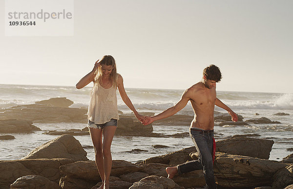 Junges Paar  das sich an den Händen hält und auf Felsen im Meer spazieren geht