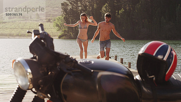 Junges Paar  das auf einem Steg am Seeufer in Richtung Motorrad läuft