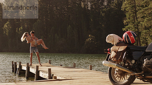 Junger Mann trägt junge Frau auf einem Steg am See in der Nähe eines Motorrads