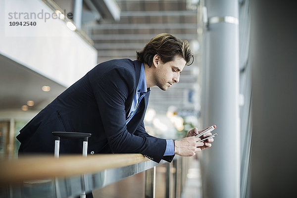 Seitenansicht des Geschäftsmannes mit dem Handy beim Anlehnen an das Geländer am Flughafen