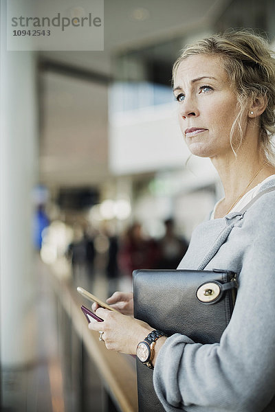 Geschäftsfrau schaut weg  während sie ein Smartphone am Flughafen hält.