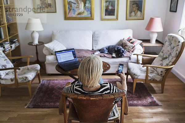 Rückansicht einer älteren Frau  die zu Hause ein Smartphone benutzt  während der Hund sich auf dem Sofa entspannt.