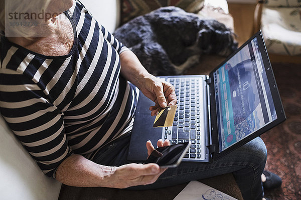Hochwinkelansicht der Seniorin mit Technik und Kreditkarte beim Sitzen mit Hund auf dem Sofa