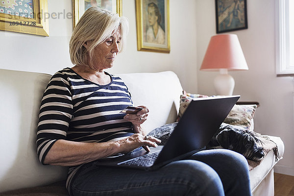 Seniorin beim Online-Shopping  während der Hund sich zu Hause auf dem Sofa entspannt