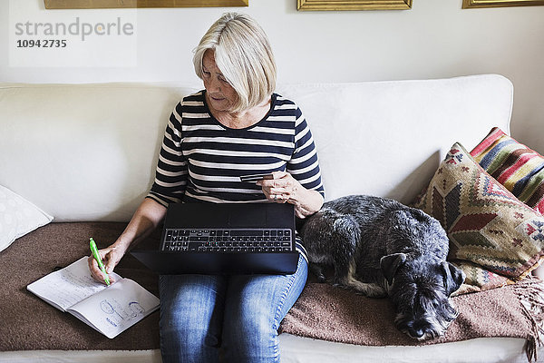 Seniorin schreibt im Buch  während sie Laptop und Kreditkarte von Hund auf dem Sofa zu Hause benutzt.