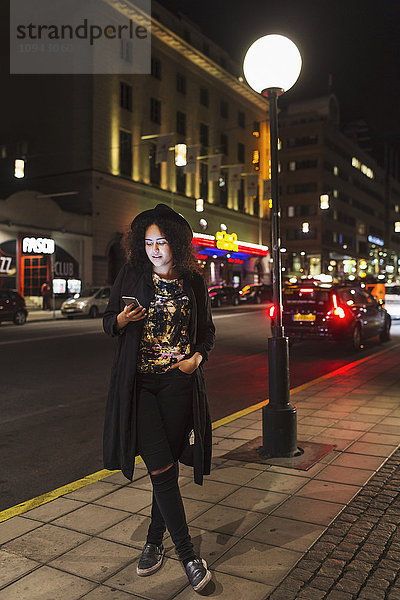 Volle Länge der Frau  die das Smartphone benutzt  während sie nachts auf dem Bürgersteig in der Stadt steht.