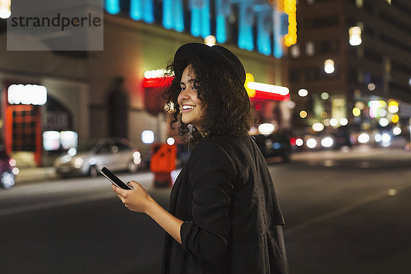 Glückliche Frau schaut weg  während sie nachts ein Smartphone in der Stadt hält.