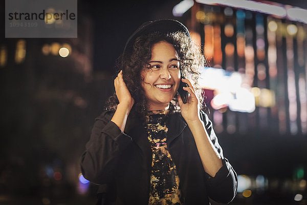 Fröhliche junge Frau schaut weg  während sie nachts in der Stadt auf das Smartphone geht.