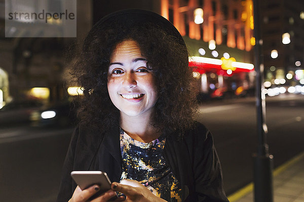 Porträt einer lächelnden Frau  die beißt  während sie nachts ein Smartphone in der Stadt hält.