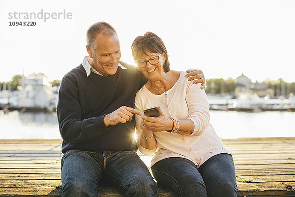 Glückliches Seniorenpaar beim Sitzen am Pier mit dem Handy
