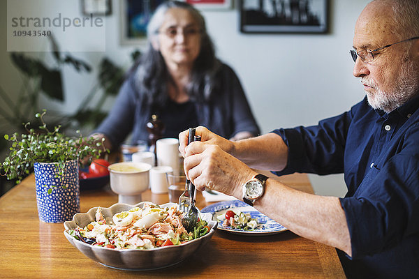 Senior Mann serviert Salat für sich selbst  während er mit einer Frau am Tisch sitzt.
