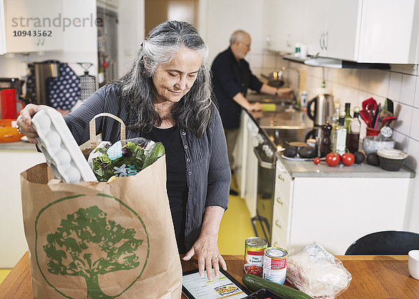 Senior Frau  die Lebensmittel aus der Einkaufstasche nimmt  während sie in der Küche ein digitales Tablett benutzt.