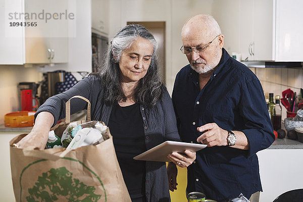 Seniorenpaar mit Einkaufstasche mit digitalem Tablett in der Küche