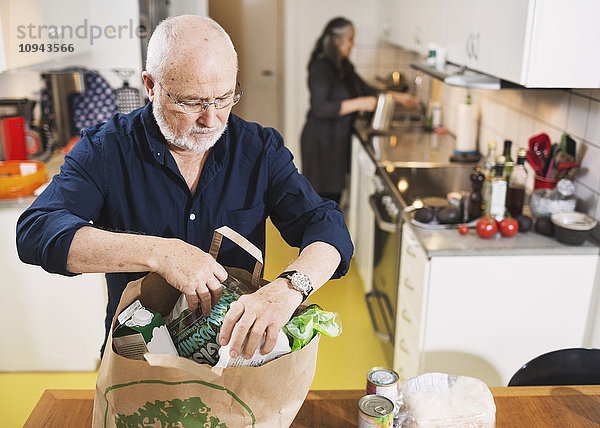 Senior Mann entfernt Lebensmittel aus der Einkaufstasche  während die Frau in der Küche arbeitet.