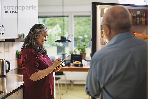 Seitenansicht einer älteren Frau  die ein digitales Tablett benutzt  während der Mann am Kühlschrank steht.
