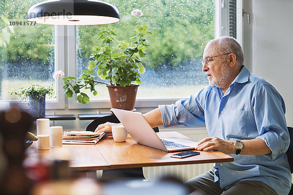 Älterer Mann mit Laptop  der nach der Zeitung am Tisch greift.