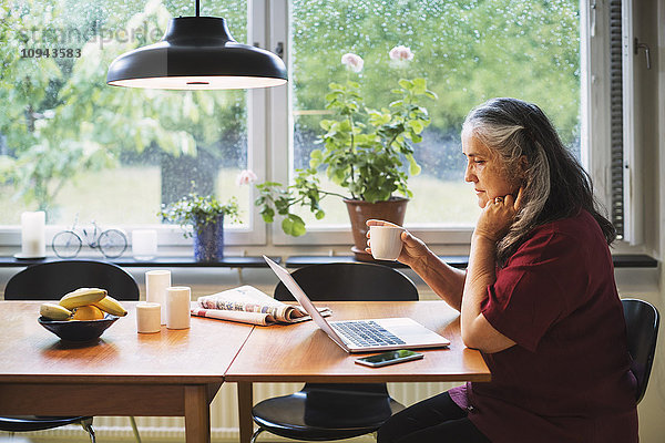 Seitenansicht der Seniorin mit Laptop beim Kaffeetrinken am Tisch