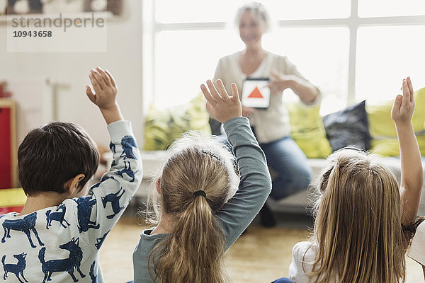 Rückansicht der Schüler  die die Hände heben  während der Lehrer das digitale Tablett im Vorschulalter zeigt.