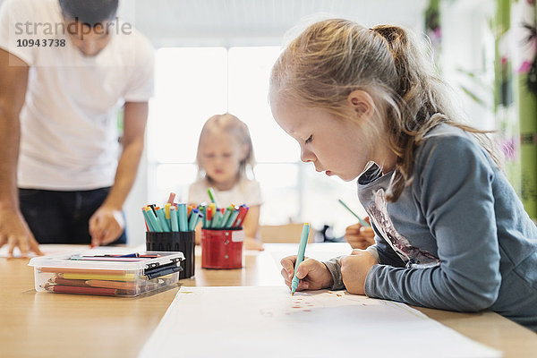 Mädchen mit Filzstift im Zeichenunterricht mit Klassenkameradin und Lehrerin im Hintergrund