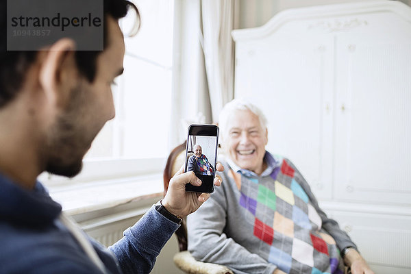 Hausmeister fotografiert glücklichen älteren Mann durch Smartphone im Pflegeheim