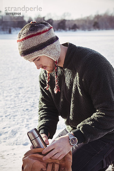 Junger Mann legt Thermoskanne in den Sack  während er auf dem schneebedeckten Feld kniet.