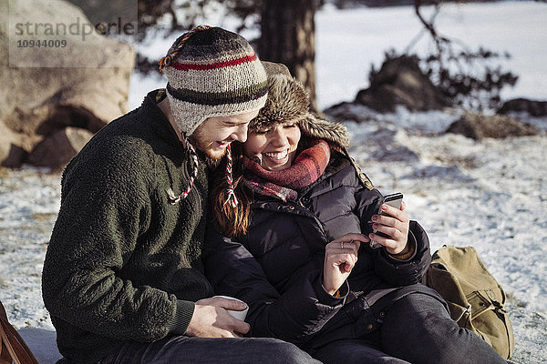 Paar mit Smartphone beim Entspannen auf schneebedecktem Feld