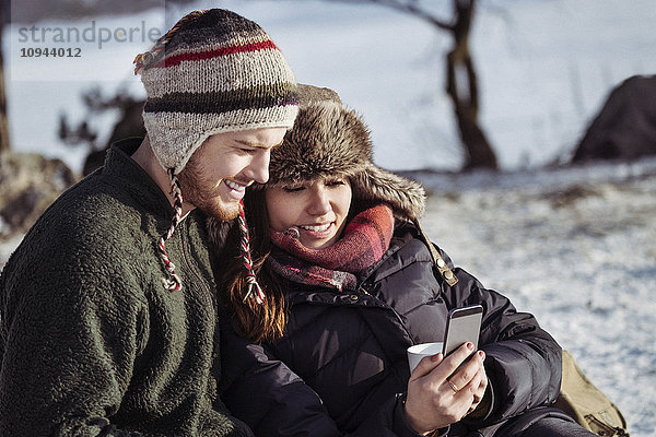 Lächelndes Paar mit Smartphone beim Entspannen auf schneebedecktem Feld