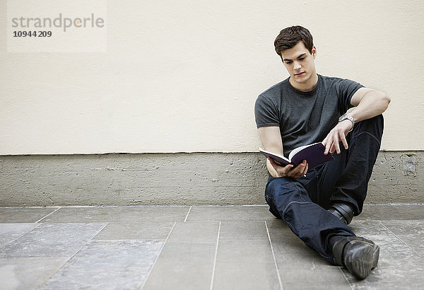 Junger Mann sitzt auf dem Boden und liest ein Buch.