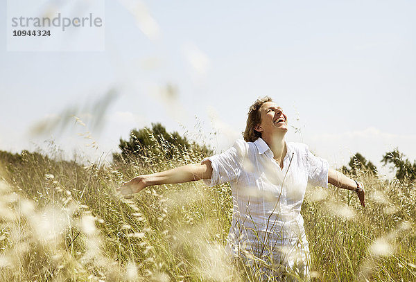 Fröhliche reife Frau lächelt beim Gehen mit erhobenen Armen im Grasfeld