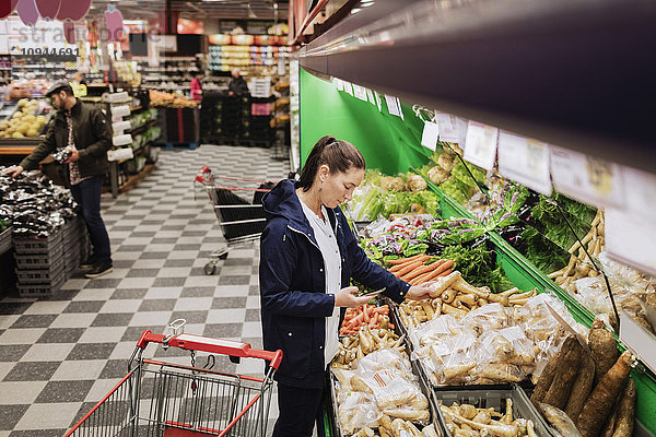 Hochwinkelansicht der Frau  die das Telefon hält  während sie im Supermarkt Gemüse kauft.