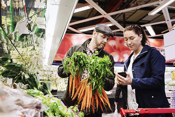 Paar mit Smartphone beim Karottenkauf im Supermarkt