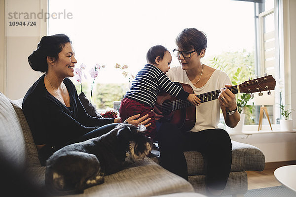 Frau mit Hund hält Kleinkind  während die Mutter zu Hause Gitarre spielt.