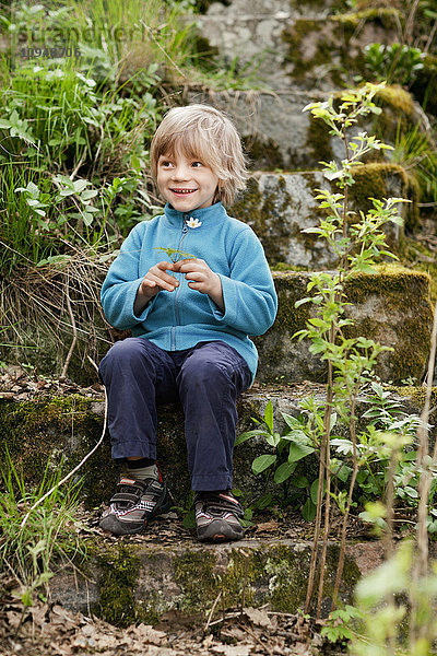Junge hält Blume und sitzt auf moosbewachsenen Stufen