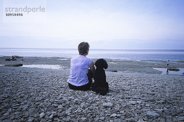 Frau und Hund sitzen am Strand und blicken auf das Wasser hinaus.
