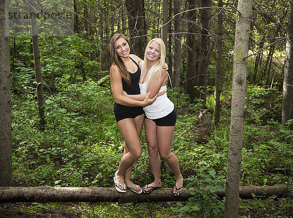 Zwei Freundinnen im Teenageralter umarmen sich und balancieren auf einem umgestürzten Baumstamm im Wald in einem Park; Edmonton  Alberta  Kanada'.