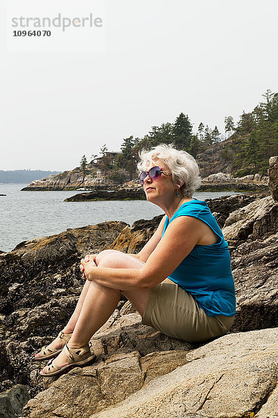 Frau sitzt auf einem Felsen am Ufer und blickt auf die Küste; Bowen Island  British Columbia  Kanada'.