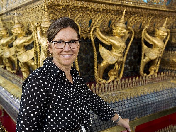 Porträt einer Frau  die neben einer Wand mit goldenen Statuen steht  Tempel des Smaragdbuddhas (Wat Phra Kaew); Bangkok  Thailand'.