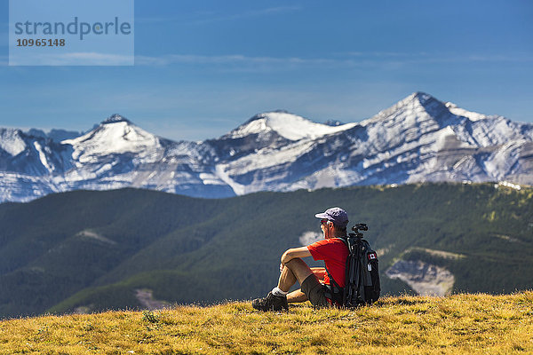 Männlicher Wanderer auf einem grasbewachsenen Berggipfel sitzend mit Blick auf Ausläufer  schneebedeckte Bergketten und blauen Himmel mit Wolken; Kananaskis Country  Alberta  Kanada