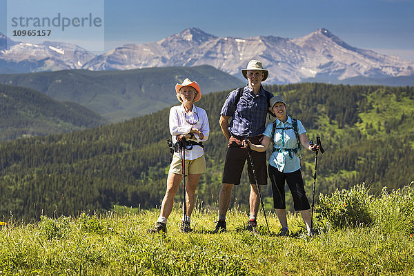 Drei Wanderer  zwei Frauen und ein Mann  auf einem grasbewachsenen Hügel mit sanften Ausläufern und einer Bergkette im Hintergrund mit blauem Himmel; Kananaskis Country  Alberta  Kanada'.