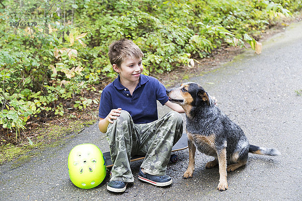 Junge  der auf einem Skateboard sitzt und seinen Hund streichelt; Salmon Arm  British Columbia  Kanada'.