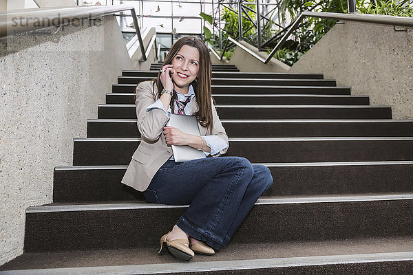 Ältere Geschäftsfrau  die mit ihrem Handy telefoniert und ein Tablet in der Hand hält  während sie auf einer Treppe im Atrium eines Bürogebäudes sitzt; Edmonton  Alberta  Kanada'.